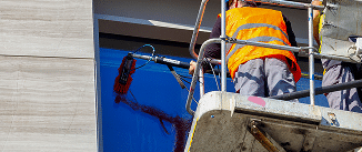 A worker wearing a safety vest operating a pressure washer on a blue exterior wall.