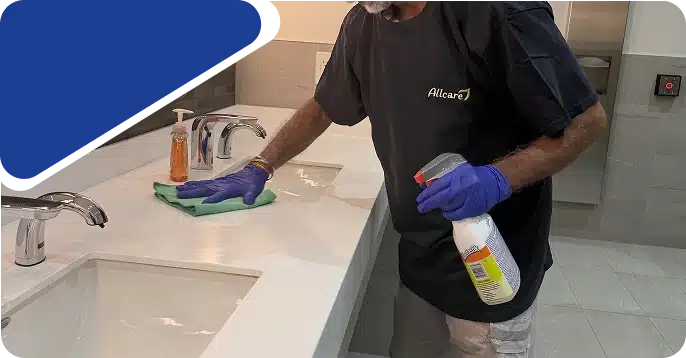 A cleaner wearing blue gloves wiping a bathroom sink counter with a spray bottle and cloth.