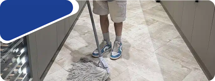 A close-up of a person's legs and a cleaning tool handle on a light-colored tiled floor.