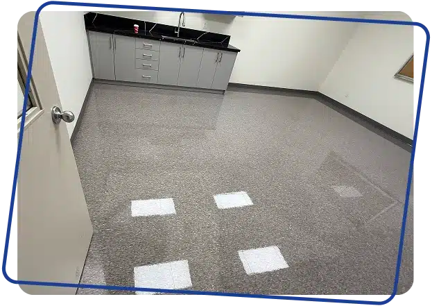 An empty room with light speckled flooring and grey cabinets under a dark countertop.