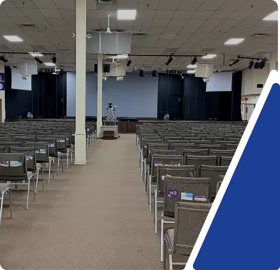 A large auditorium or place of worship with rows of chairs facing a stage area, framed by a blue graphic.