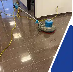 A blue floor buffing machine sitting on a wet tiled floor with a yellow power cord.